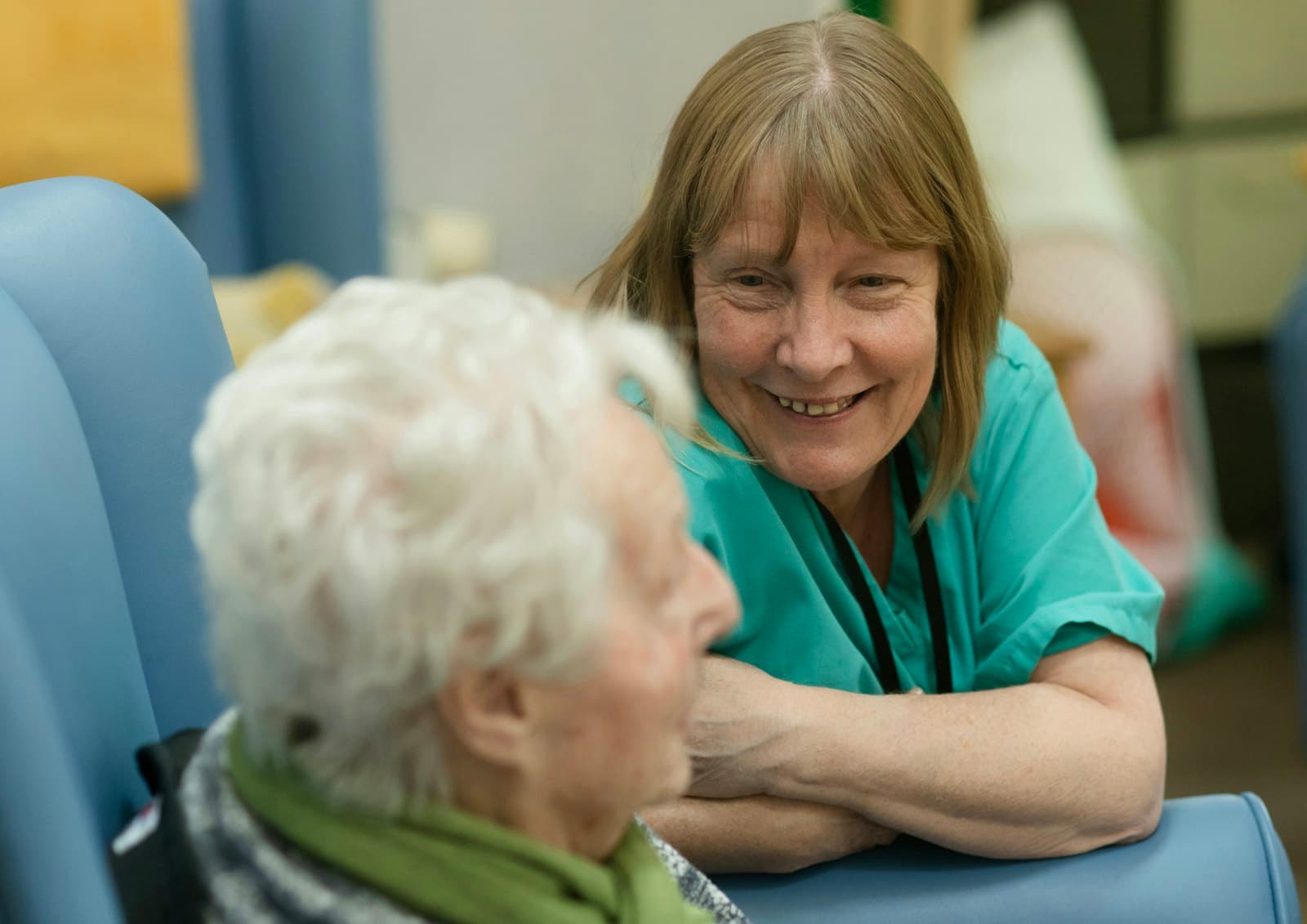 A caregiver in green scrubs smiling with an older woman in her chair